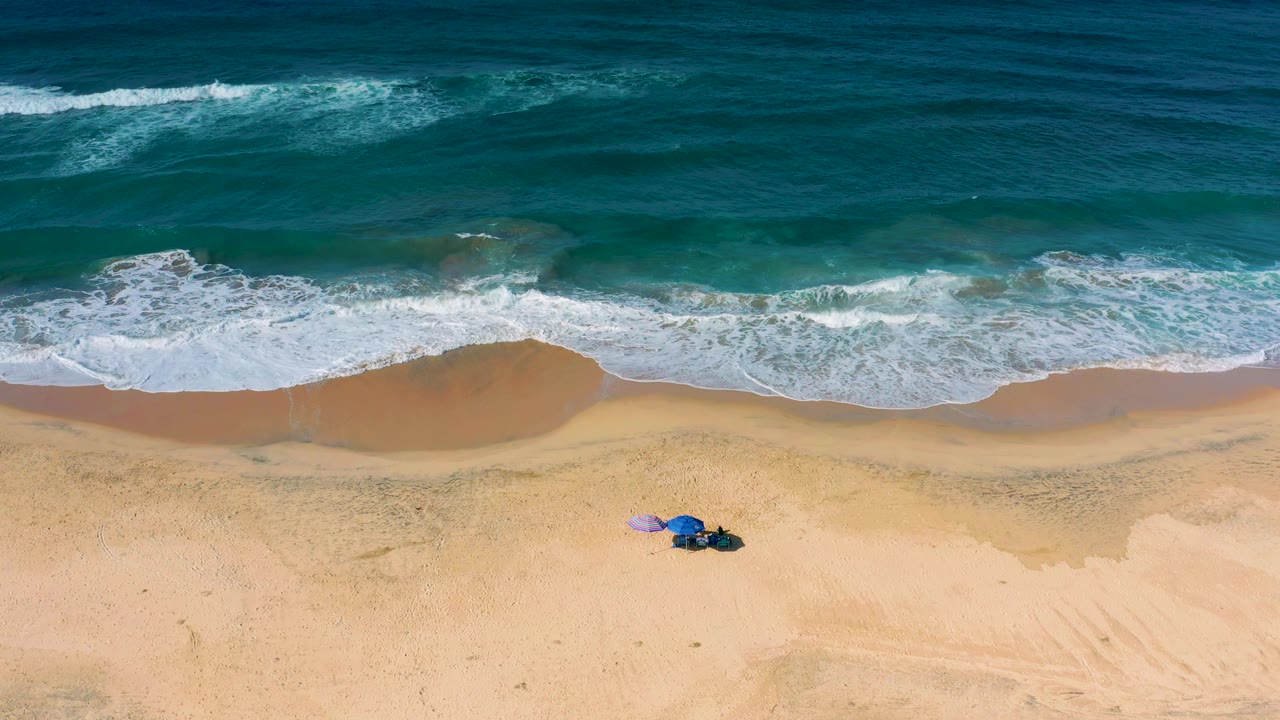 At mid-clip, white water curls along the Cerritos shore while the lower sand shows faint tracks and a small colored item cluster, with no visible people.