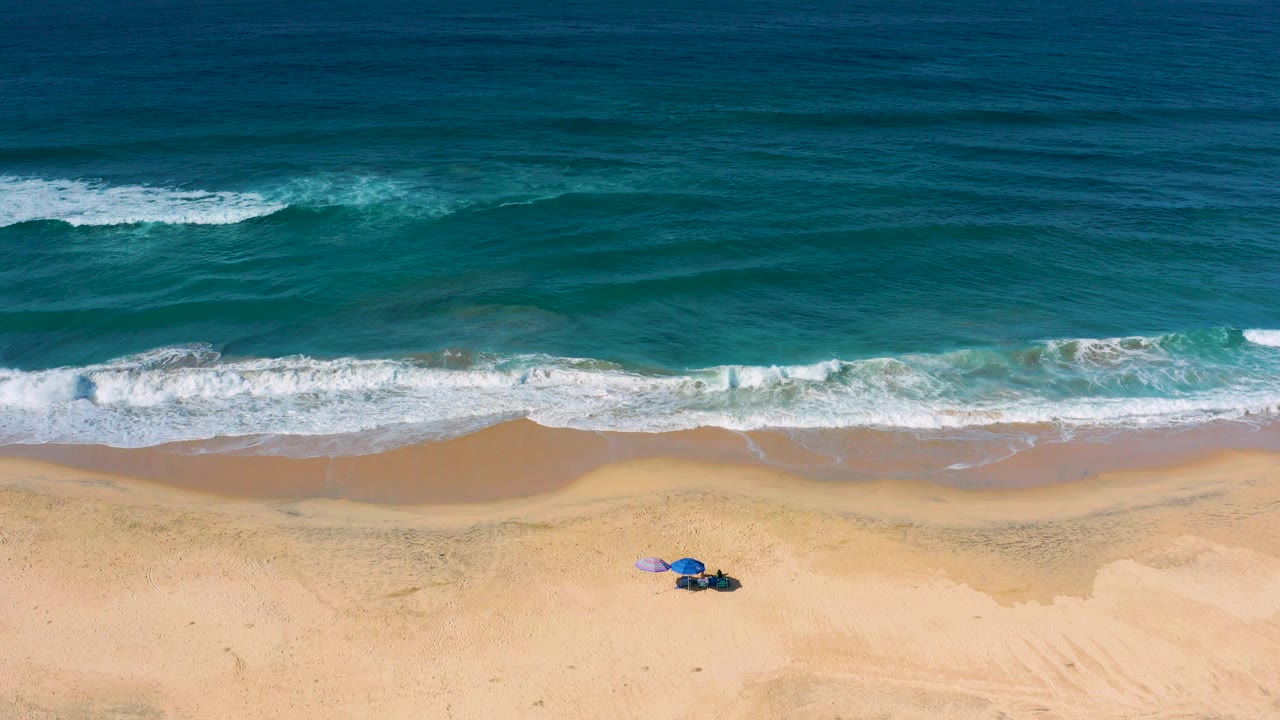 Aerial frame from Cerritos, Baja California Sur, Mexico showing blue ocean, white surf, sandy beach, and a small blue umbrella setup on the sand with no visible people.