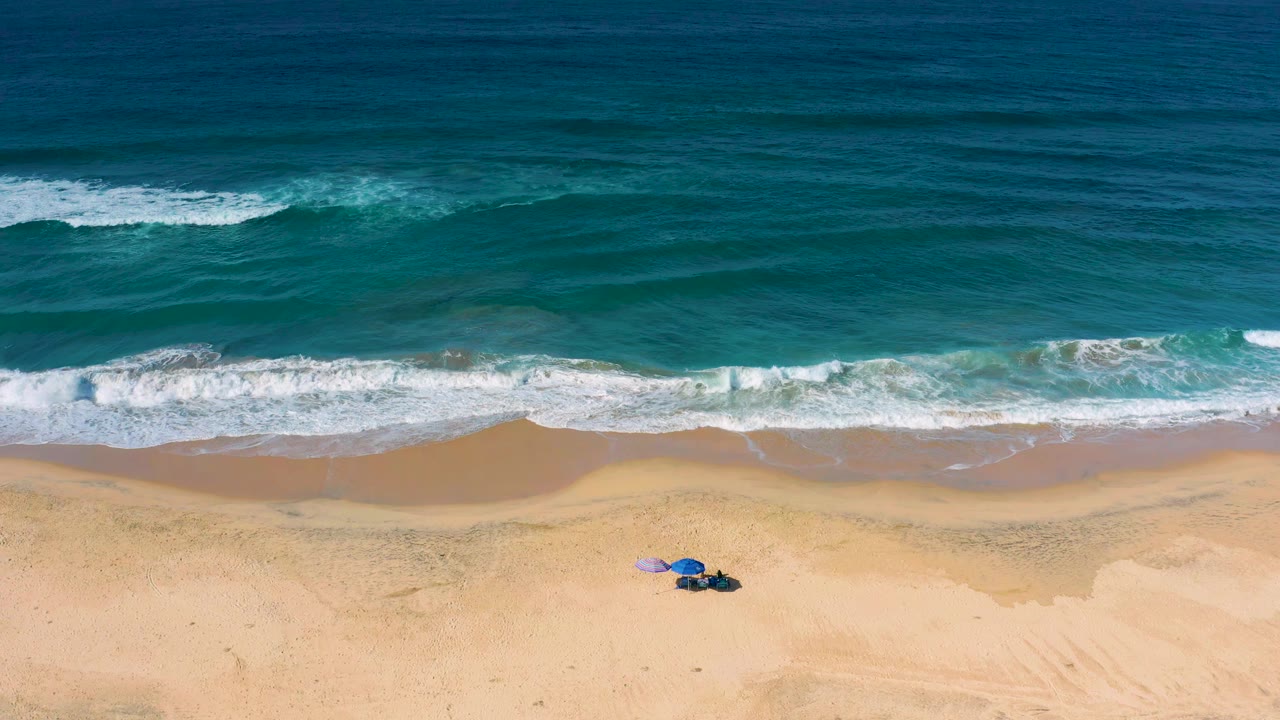 Second frame over Cerritos beach with the white wave line slightly farther across the shore, deep blue water above, pale sand below, and no visible people.