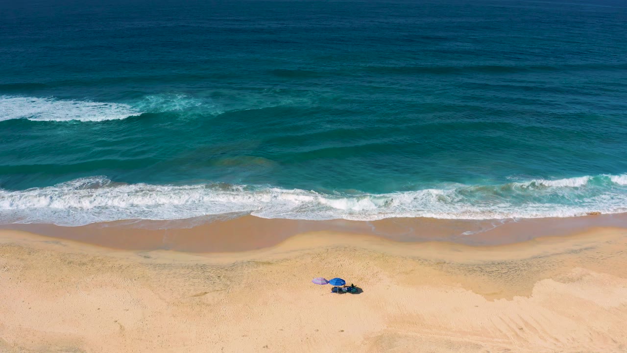 Aerial Cerritos beach frame with layered teal and dark blue water, broken white waves near shore, a broad sand strip, and no visible people around the small umbrella setup.