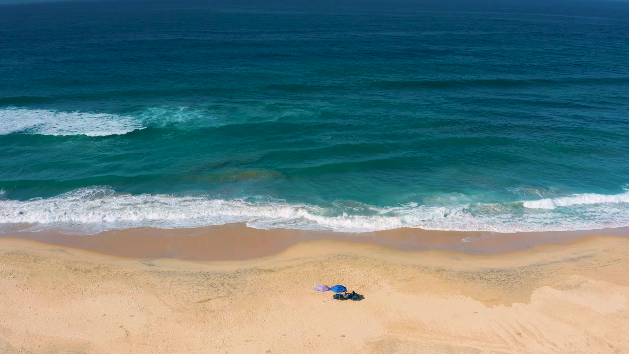 Cerritos shoreline frame showing foam spread across the wet sand, open blue water beyond, a small blue umbrella setup on dry sand, and no visible people.