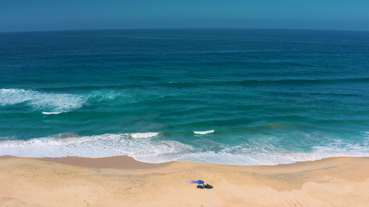 Opening aerial frame over Cerritos, Baja California Sur, with deep blue water, white surf, pale sand, small umbrellas near the lower edge, and no visible people.