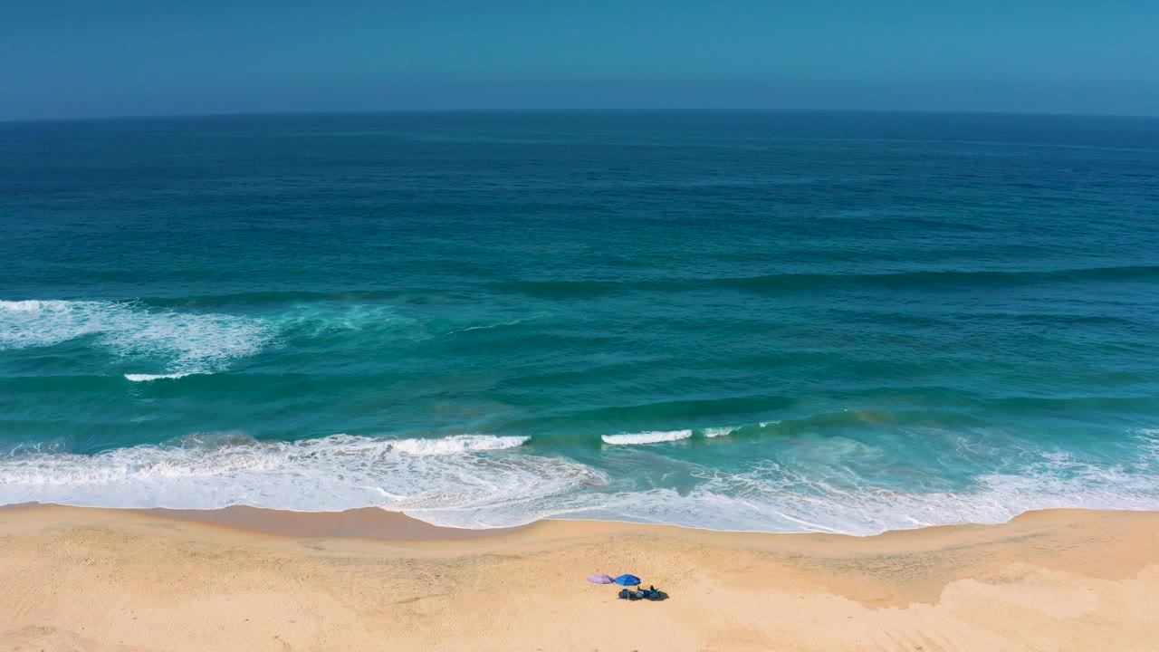 At 0:02 the Cerritos shoreline remains in a high aerial composition, with broken white wave patches across teal water and a small umbrella cluster low on the sand, no visible people.