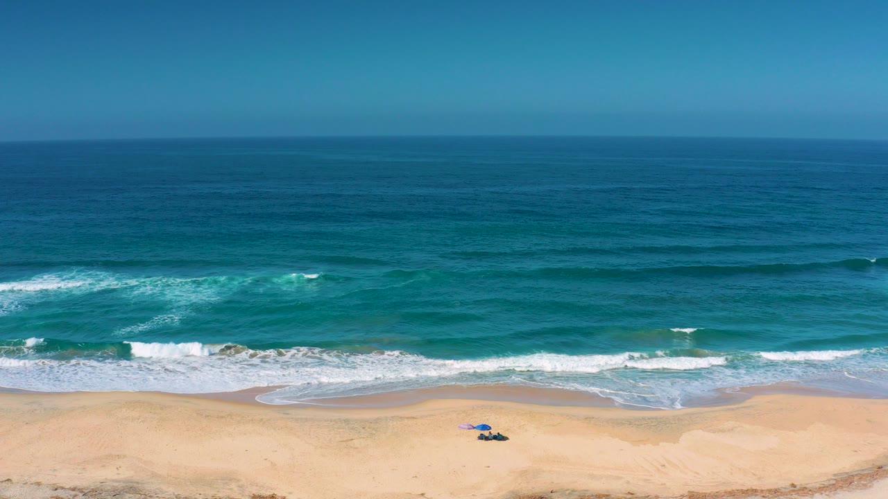 Wide aerial view of Cerritos, Baja California Sur, with blue ocean bands, white surf reaching the sand, a small blue beach setup near the lower edge, and no visible people.