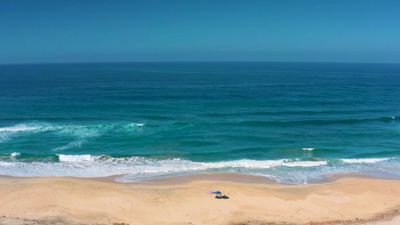 Aerial beach frame at Cerritos showing a white foam line curling across the near shore, turquoise water beyond it, dry sand below, and no visible people.
