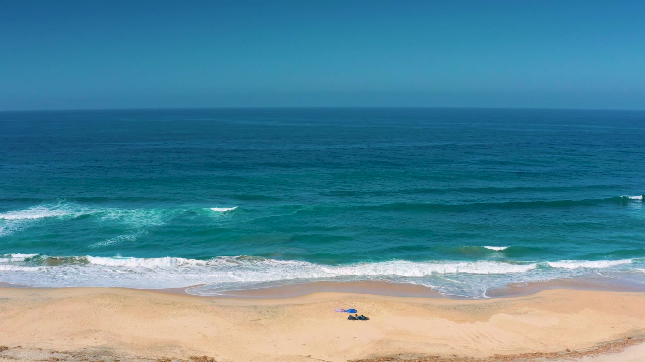 Elevated view of Cerritos beach with several low wave bands, white foam near the shoreline, a small blue beach setup on the sand, and no visible people.