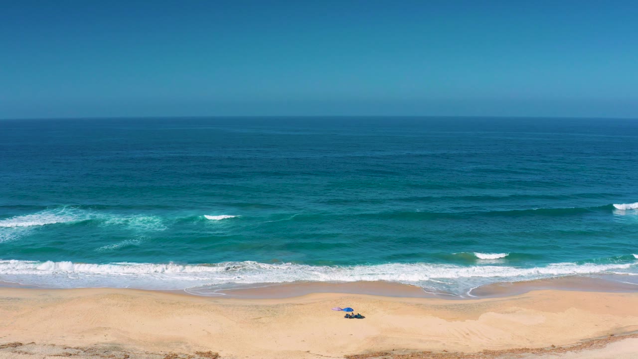 Cerritos shoreline seen from above with a broad blue ocean field, thin white breaks, pale sand along the bottom, and no visible people.