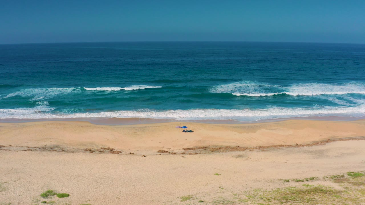 Aerial view of Cerritos, Baja California Sur, Mexico, with blue ocean waves rolling toward broad empty sand, a small blue beach setup near the waterline, and no visible people.
