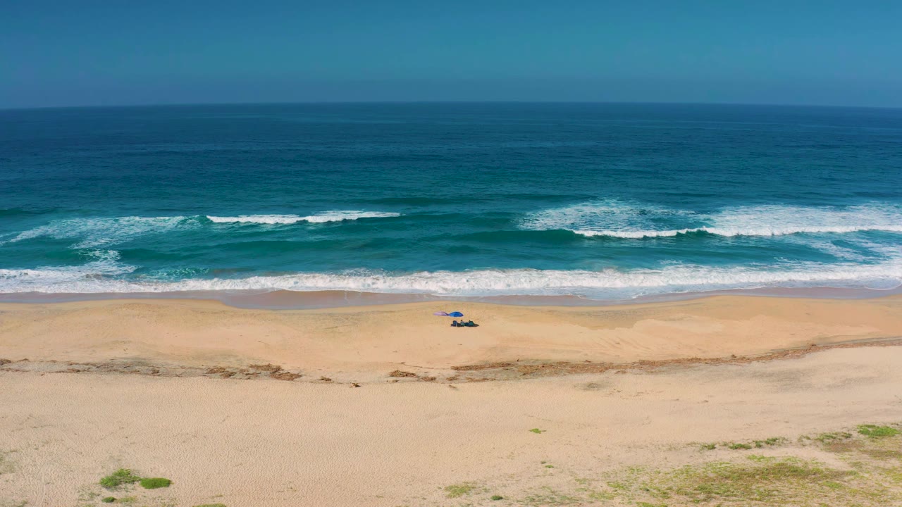 Aerial frame over Cerritos beach showing white surf spreading across blue water, open sand in the foreground, a small blue object near the shore, and no visible people.
