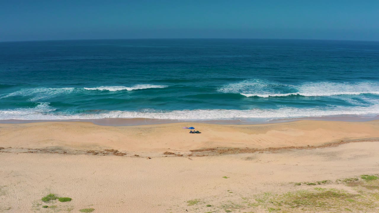 Cerritos aerial beach frame with parallel white wave bands, turquoise and deep blue water, broad tan sand, a small blue beach setup near the shore, and no visible people.