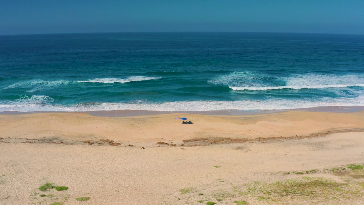 Aerial view in Cerritos with foamy waves close to the shoreline, a wide quiet beach below, a small blue beach setup near the water, and no visible people.