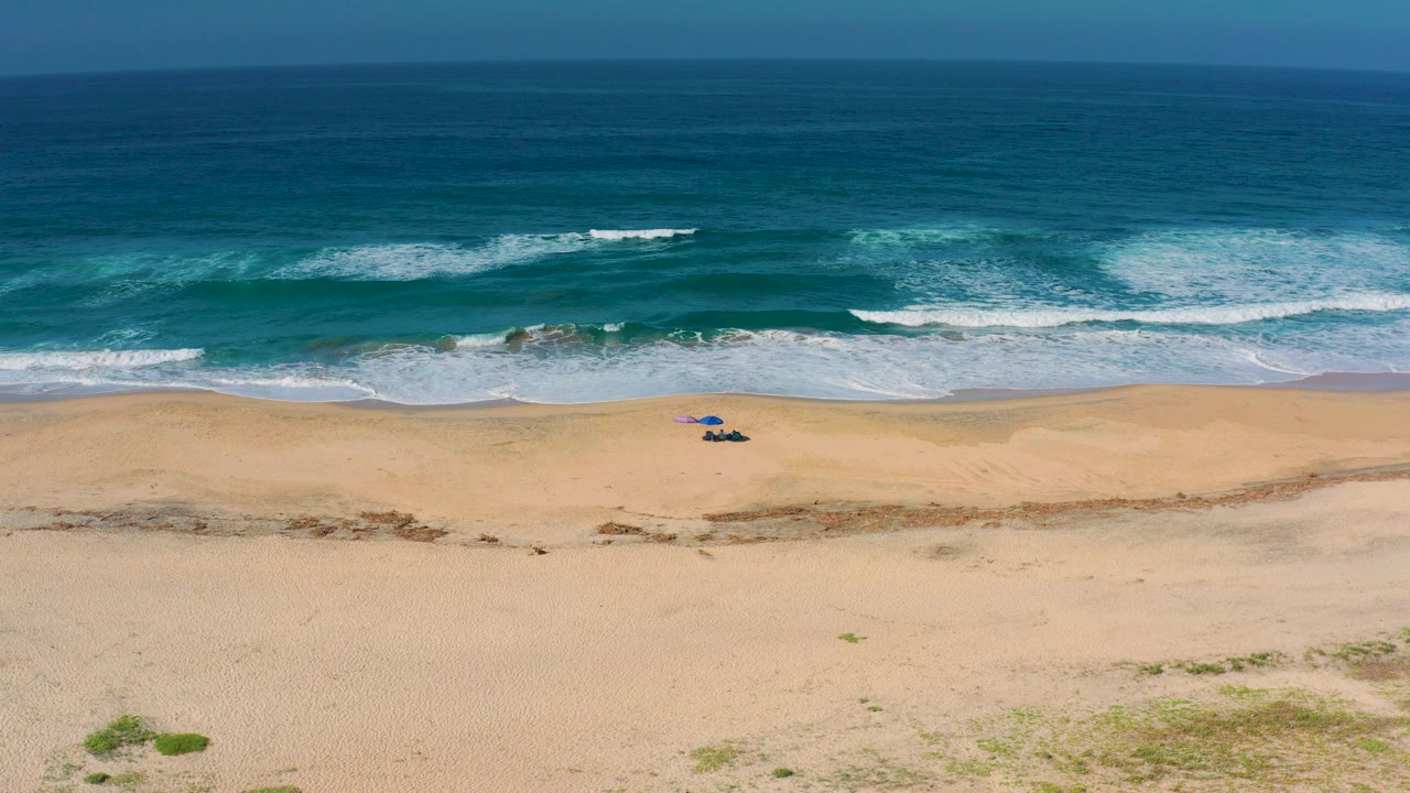Aerial view of Cerritos beach in Baja California Sur with blue-green ocean, white surf bands, open tan sand, and tiny people near a small umbrella setup.