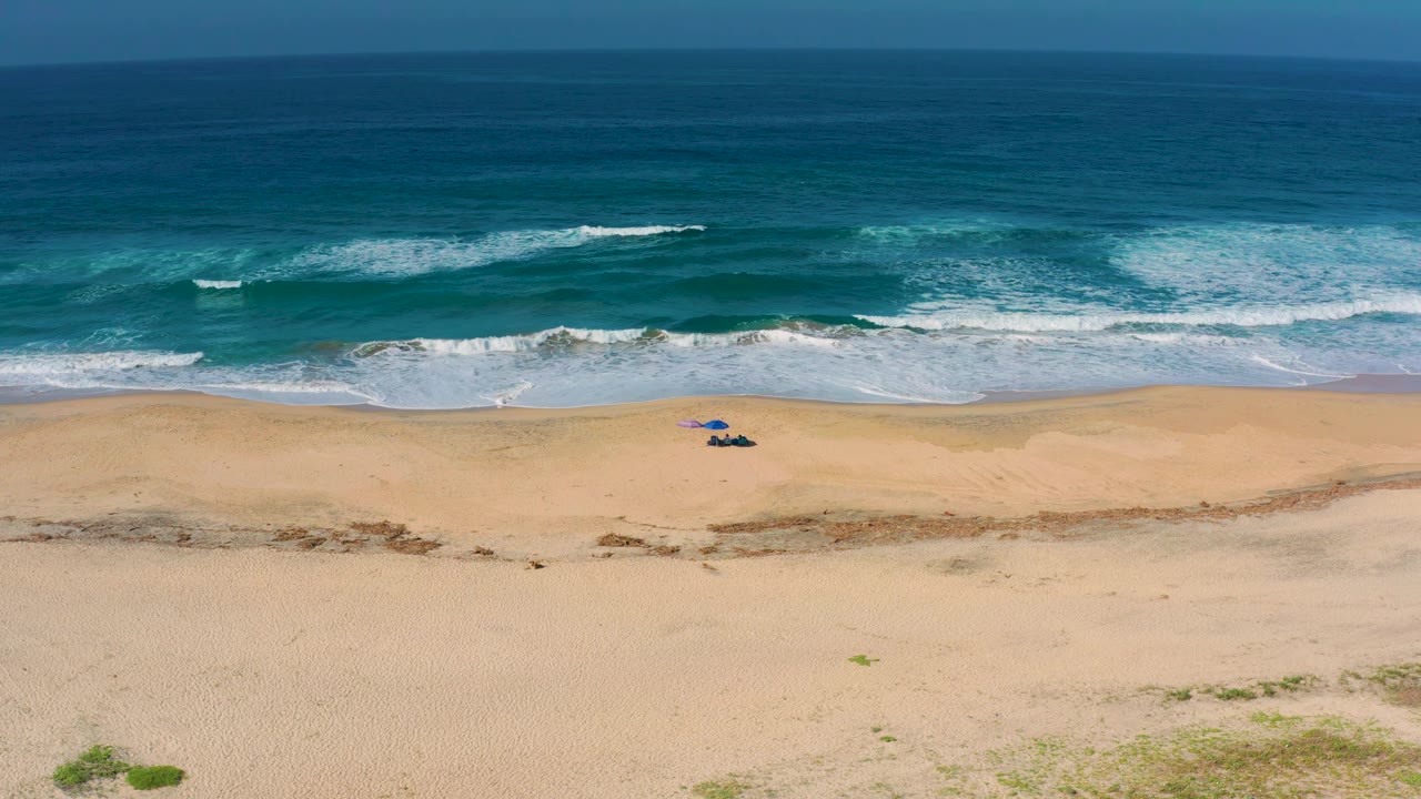 High view over Cerritos beach with layered turquoise water, foamy breakers, broad sand, and a compact beach setup set away from the shoreline.