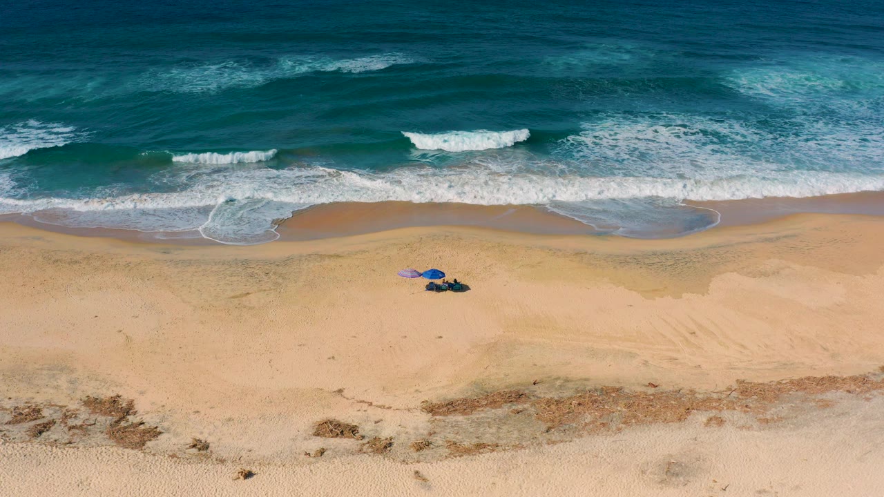 Overhead frame of Cerritos beach with teal surf at the top, tan sand below, and small umbrellas with tiny visible people near the lower left of the beach.
