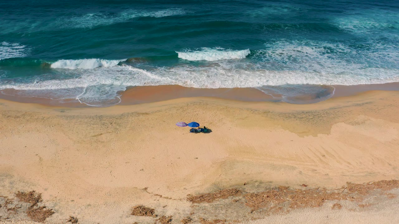 Drone-style overhead frame of Cerritos with darker teal water, a foamy shore break, broad tan sand, and the umbrella setup with tiny visible people near the lower center.