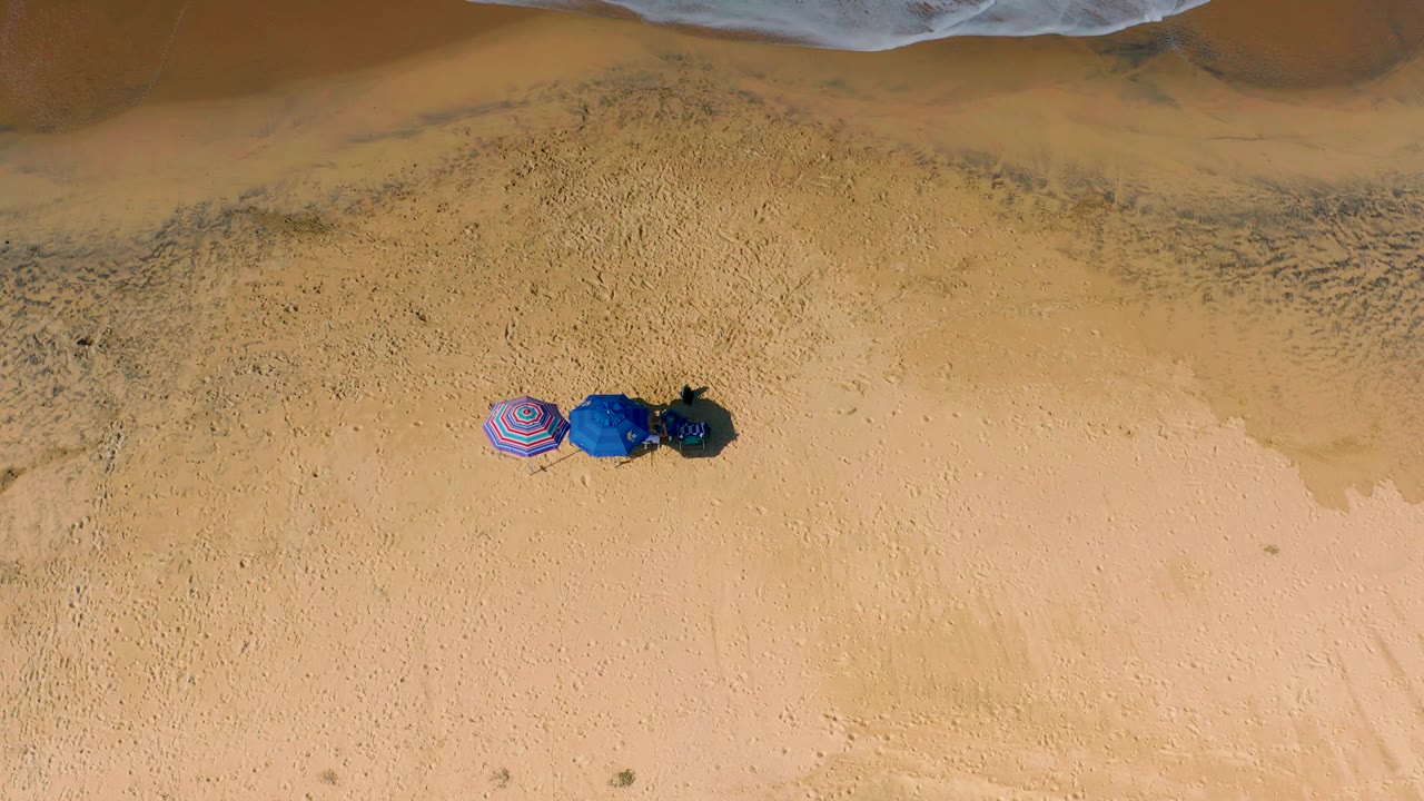 Top-down Cerritos beach view with three umbrellas near the upper middle of the sand, pale surf foam along the top border, and no visible people.