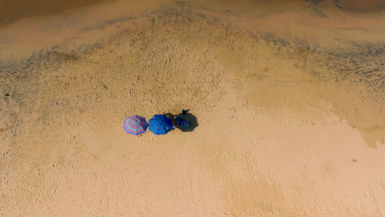 Aerial view over Cerritos sand with the three umbrellas lower left of center, mottled wet sand above them, and no visible people.