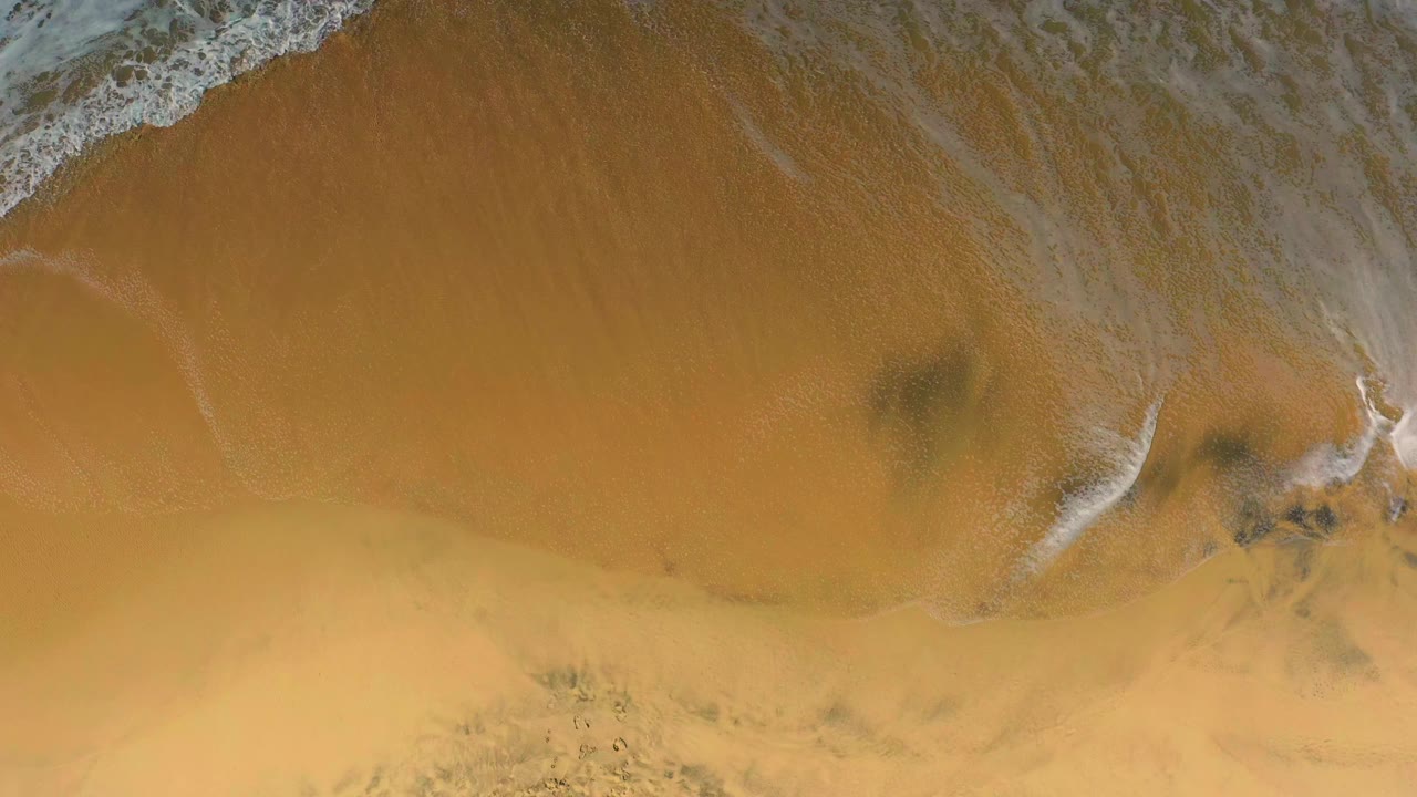 Aerial frame of Cerritos beach sand with the foam line sliding along the top edge, faint water streaks, darker marks, and no visible people.