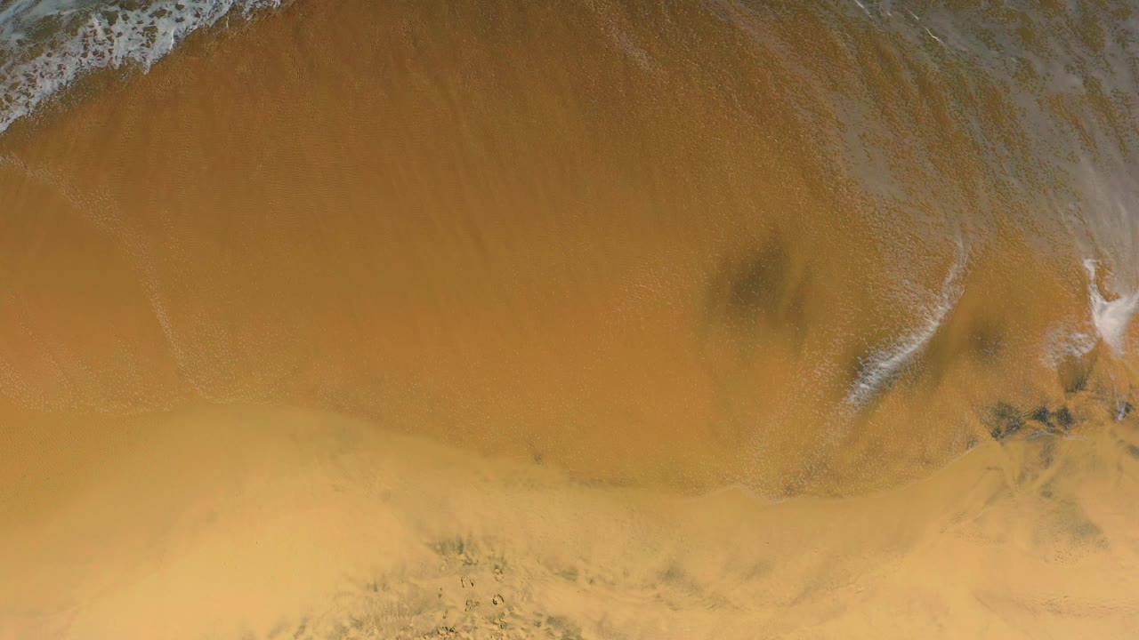 Overhead Cerritos shoreline frame with a curved white surf edge, smooth orange tan sand below it, small darker beach markings, and no visible people.