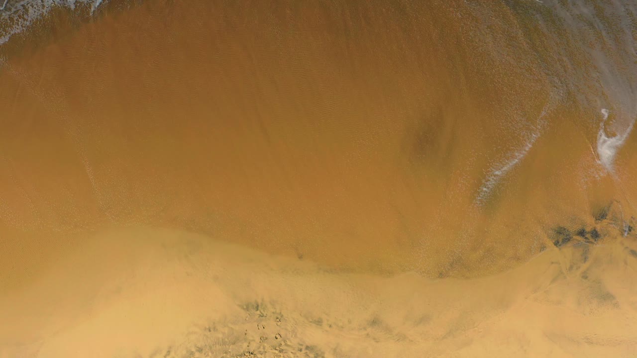 Aerial view over Cerritos beach where most of the frame is tan sand, with thin foam streaks near the upper edge, darker wet areas, and no visible people.