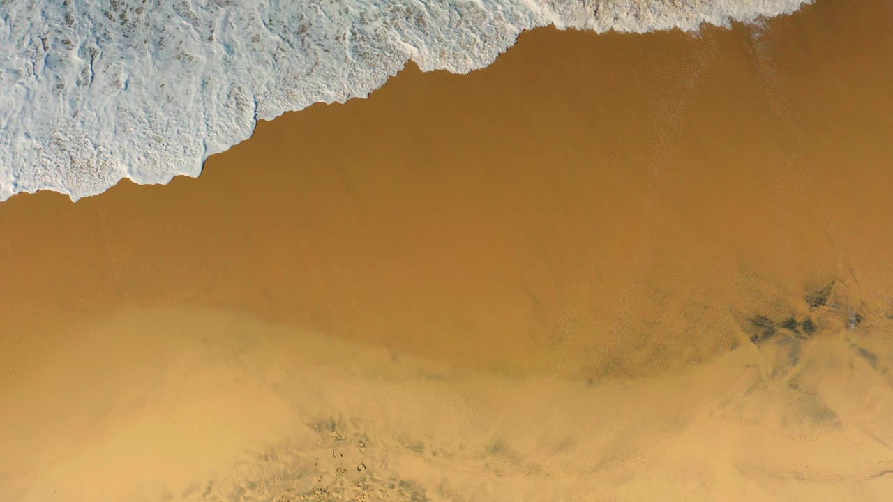 Overhead view of white surf entering from the top edge over golden sand at Cerritos, Baja California Sur, with darker shallow-water patches and no visible people.