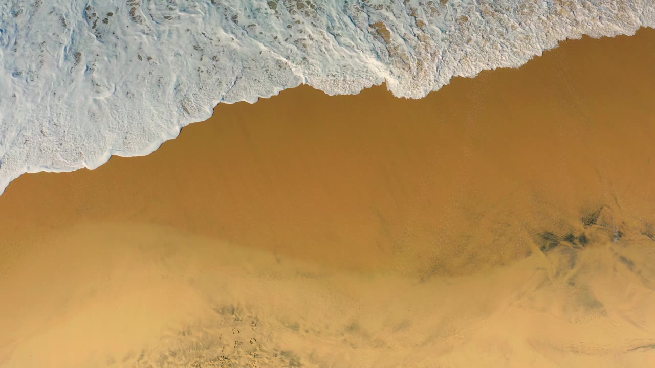 A wider band of surf foam covers the upper third of the Cerritos beach frame while tan sand and faint dark patches remain visible below, with no visible people.