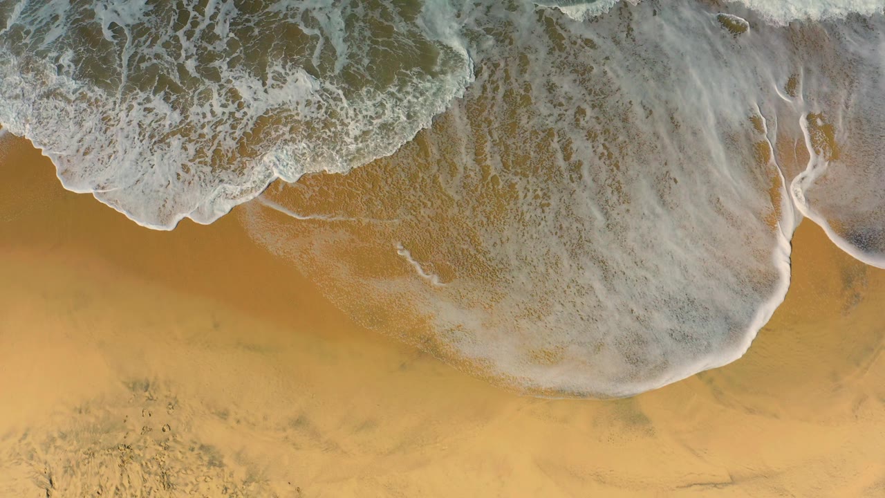White surf foam pulls across smooth golden sand at Cerritos, leaving pale water films and darker wet patches, with no visible people.
