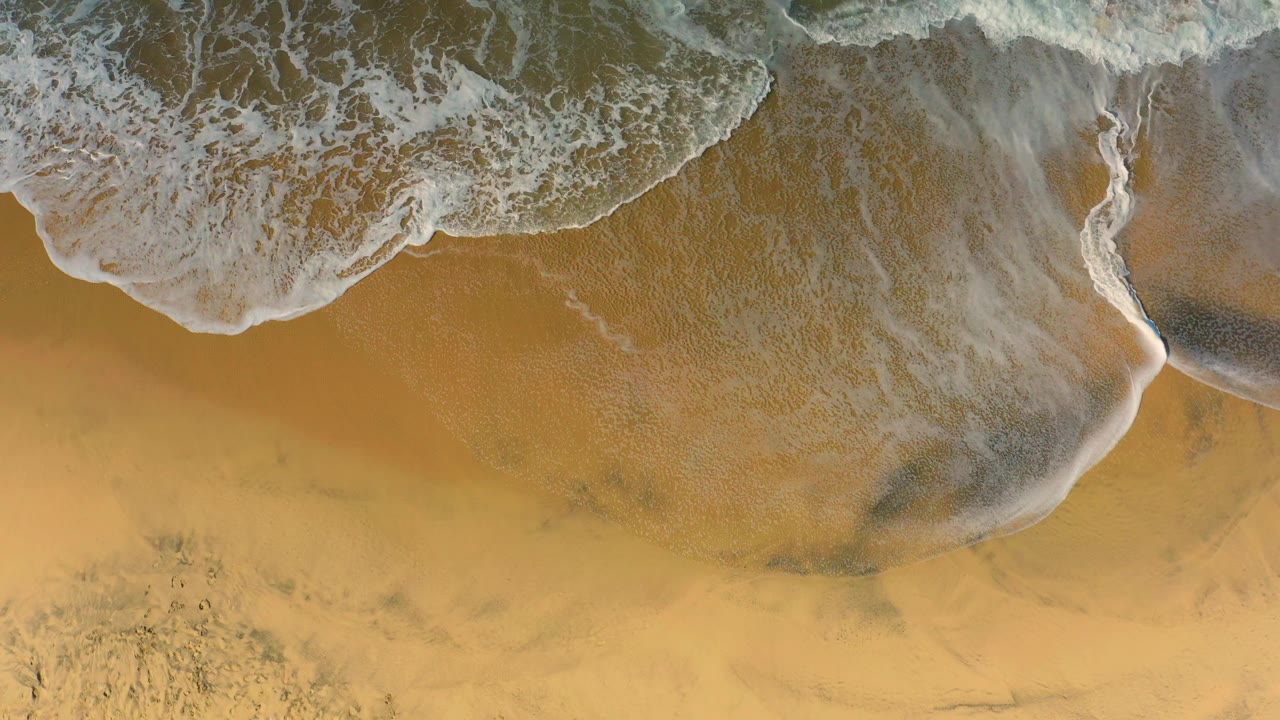 A diagonal wash of white foam spreads from the upper left over warm Cerritos sand, with translucent water and no visible people.