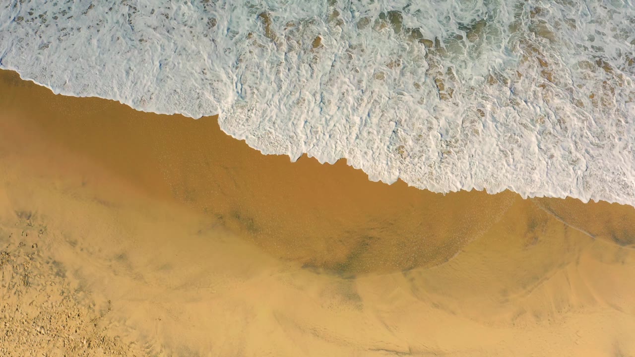 Thick white foam occupies the top of the frame while smooth golden sand remains visible below at Cerritos, Baja California Sur, with no visible people.