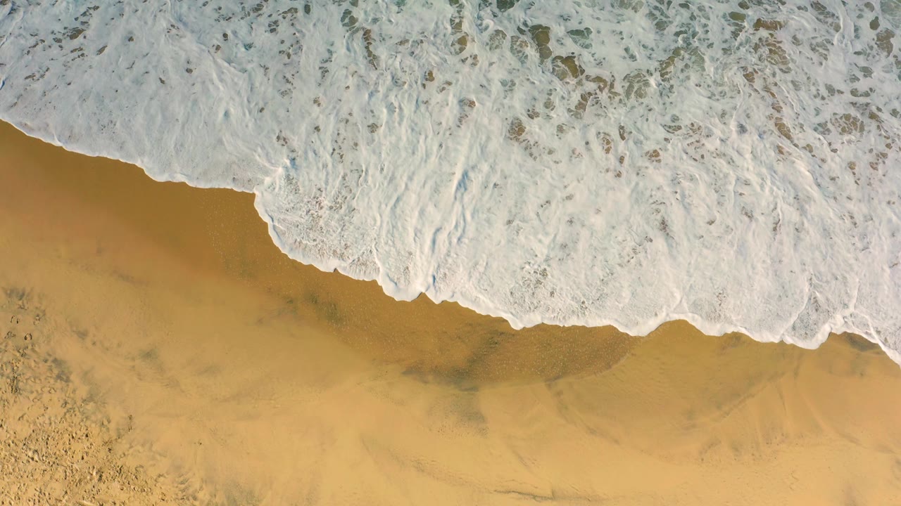 A broad diagonal sheet of white surf advances over empty golden sand in Cerritos, Baja California Sur, with no visible people.