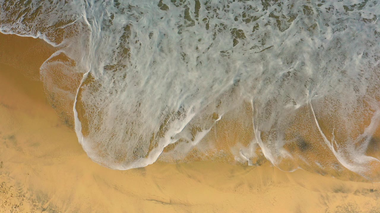 Overhead view of white foam spreading over golden sand at Cerritos, Baja California Sur, with dark wet patches and no visible people.