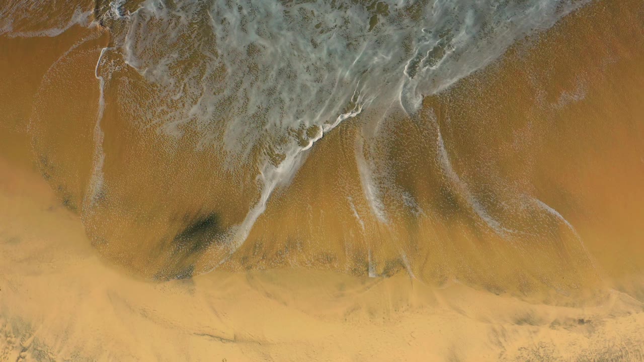 An overhead beach frame shows foam thinning across darker wet sand patches and golden sand at Cerritos, with no visible people.