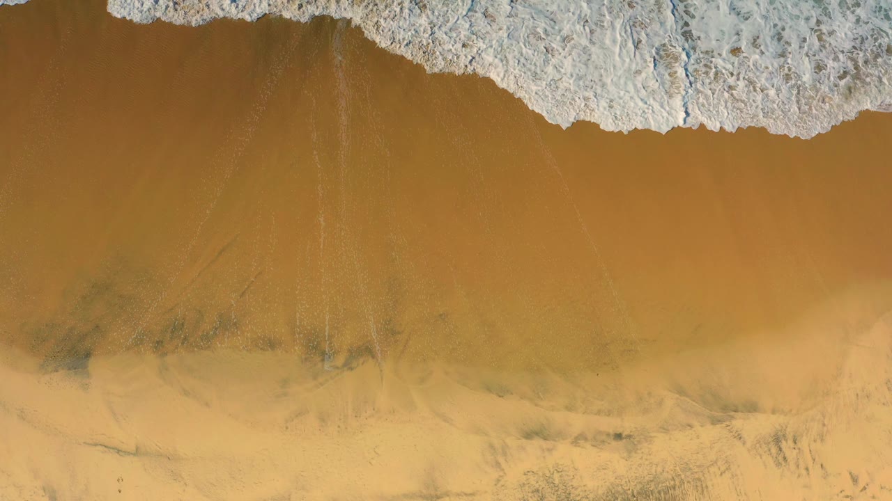 An overhead frame shows a curved band of white surf foam above amber wet sand in Cerritos, Baja California Sur, with no visible people.