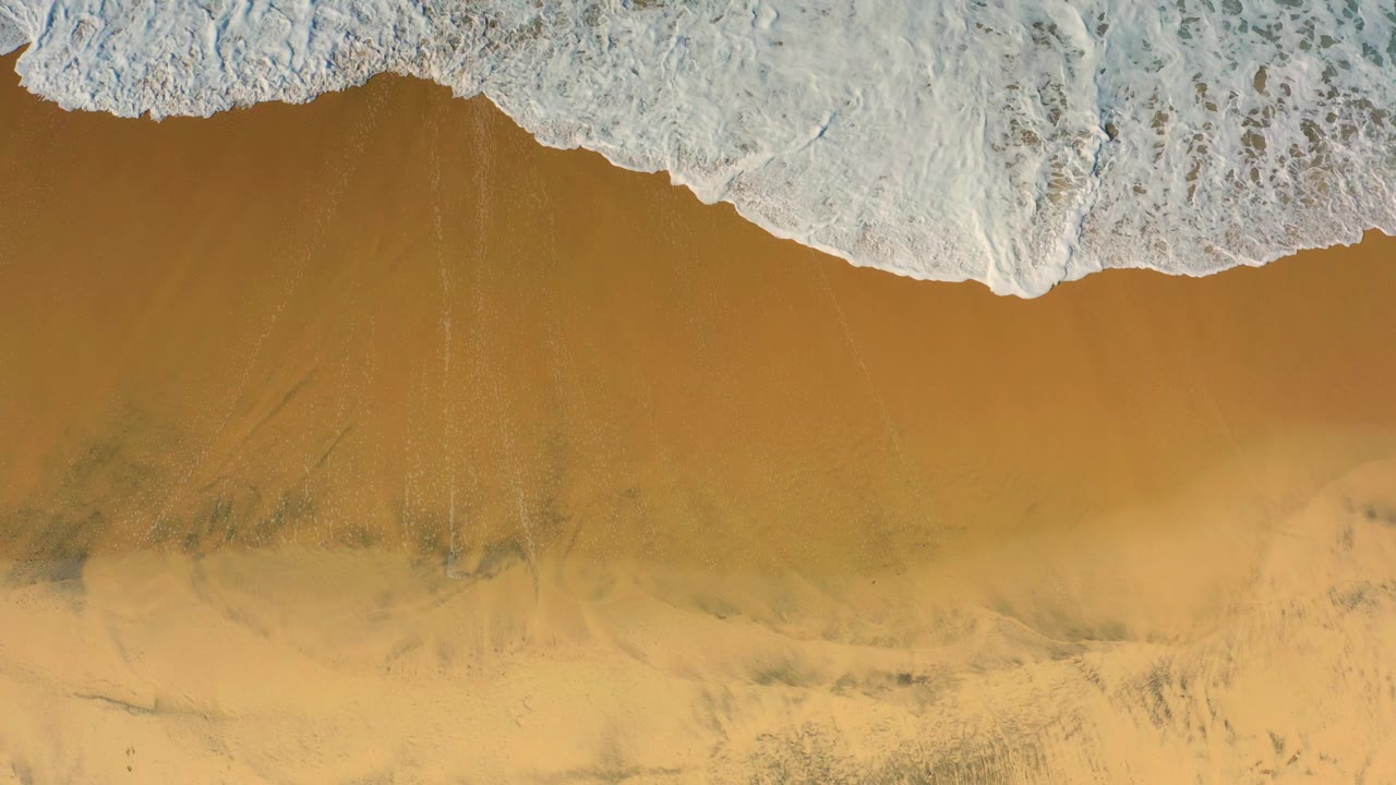 White surf foam advances deeper into the top half of the overhead Cerritos beach frame, leaving golden sand visible below and no visible people.