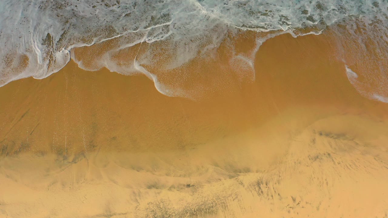 Aerial frame of thin white surf curling over darker wet sand at Cerritos beach, with no visible people.