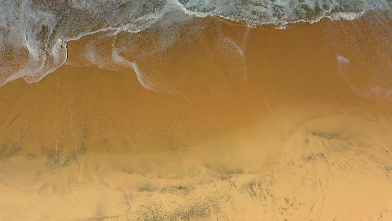 Overhead Cerritos shoreline detail with white wave foam pressing into smooth amber sand, with no visible people.