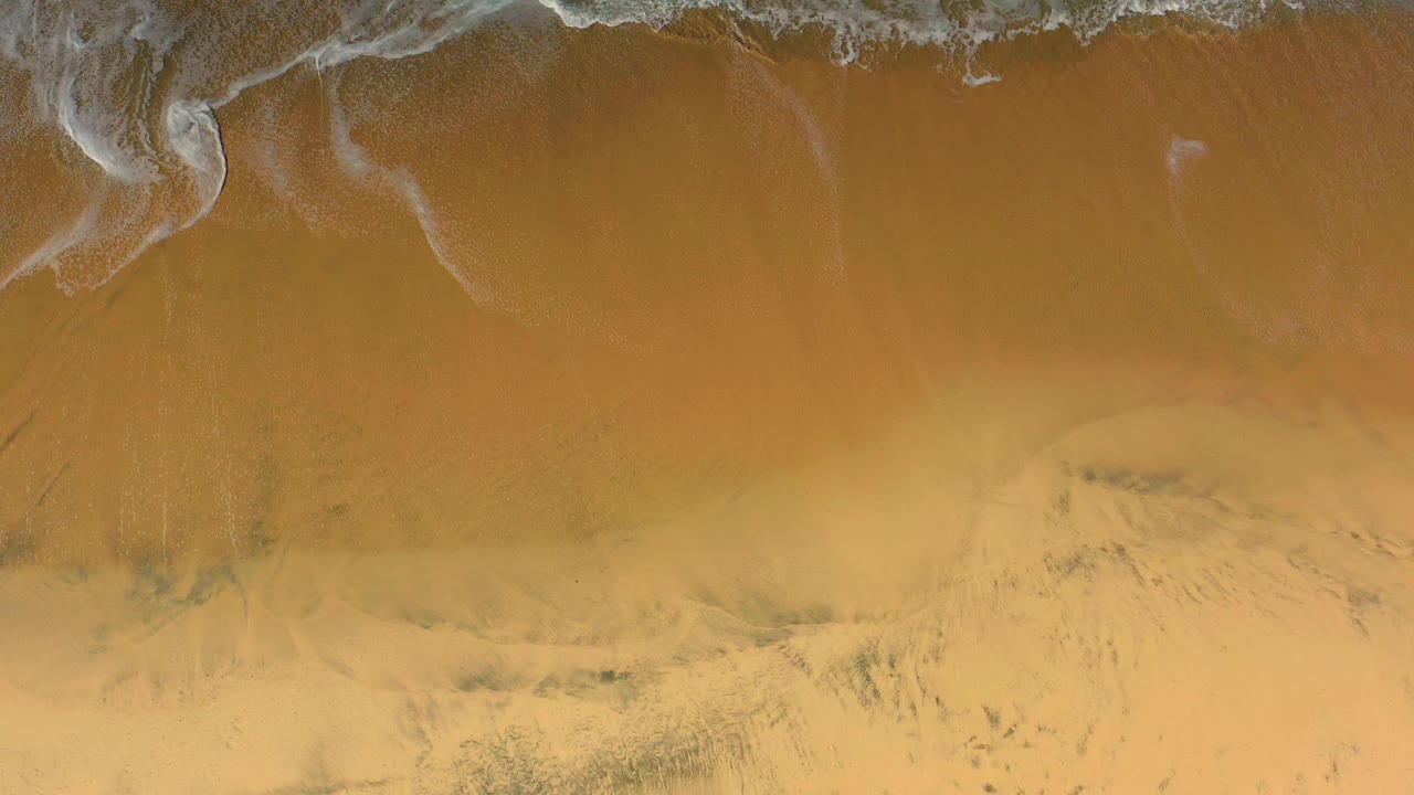 Aerial view of faded white foam traces over empty golden Cerritos beach sand, with no visible people.