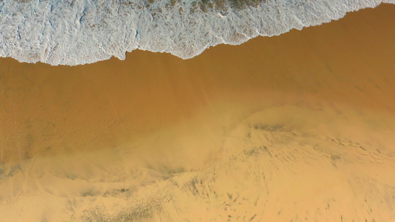 Overhead view of white foam touching golden Cerritos beach sand at the start of the clip, with no visible people.