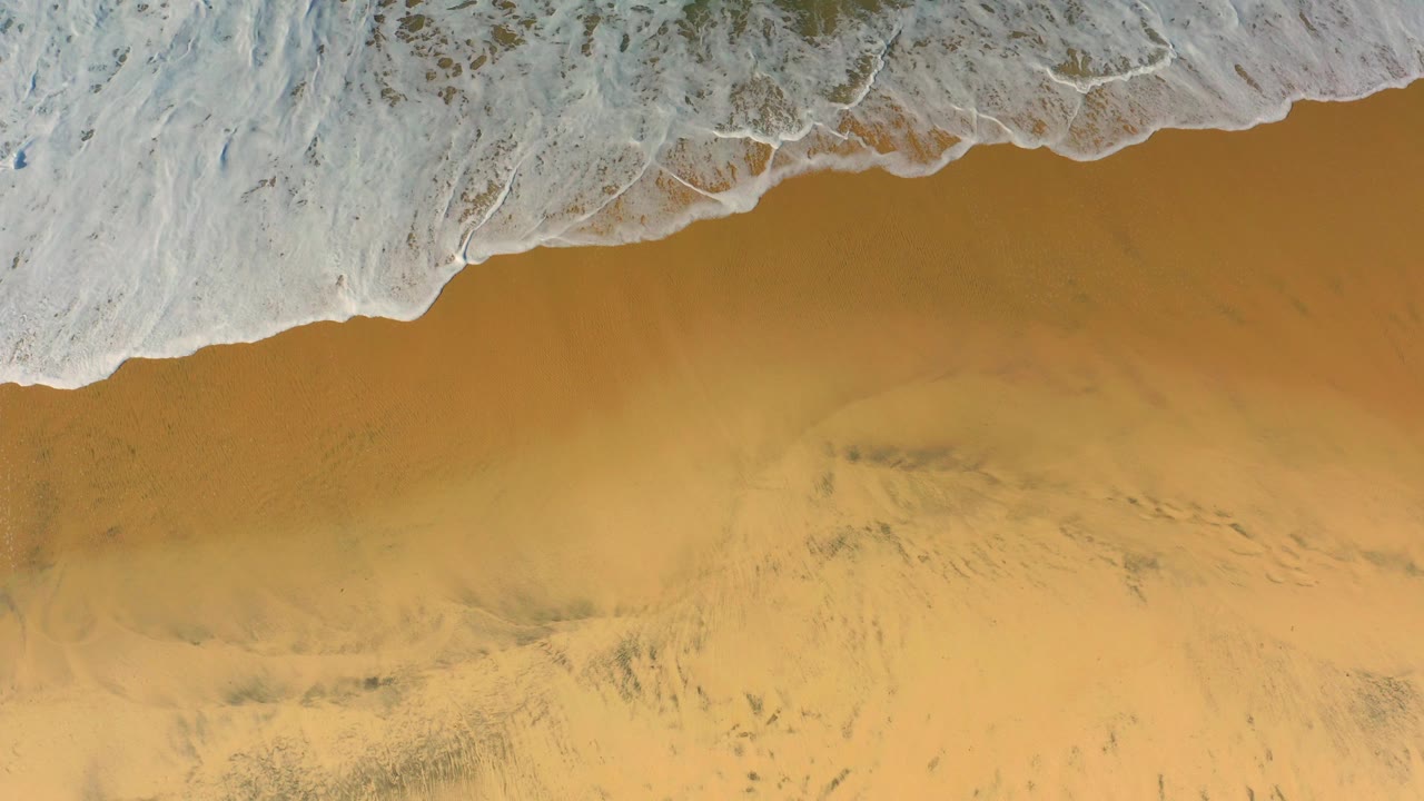 Foam spreads farther across the upper left of the Cerritos beach frame while the lower area remains open golden sand, with no visible people.
