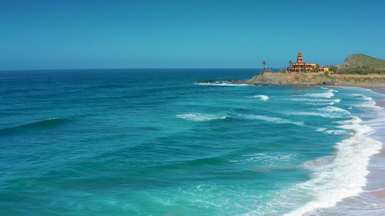 At 0:02, the frame holds a wider ocean surface with bands of white foam approaching the right shoreline and the Cerritos bluff buildings near the upper right, with no visible people.