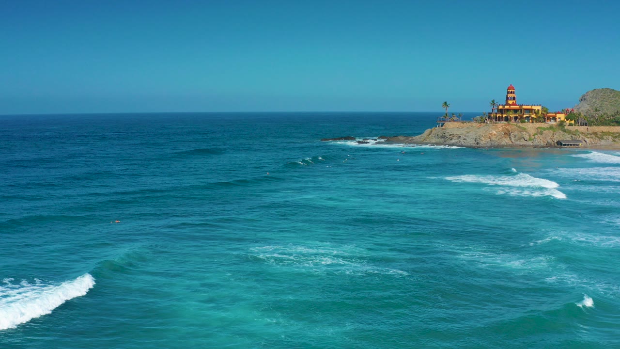 Wide aerial view of Cerritos, Baja California Sur with turquoise water, white surf, a rocky point, a warm-toned building at upper right, and tiny distant people in the water.
