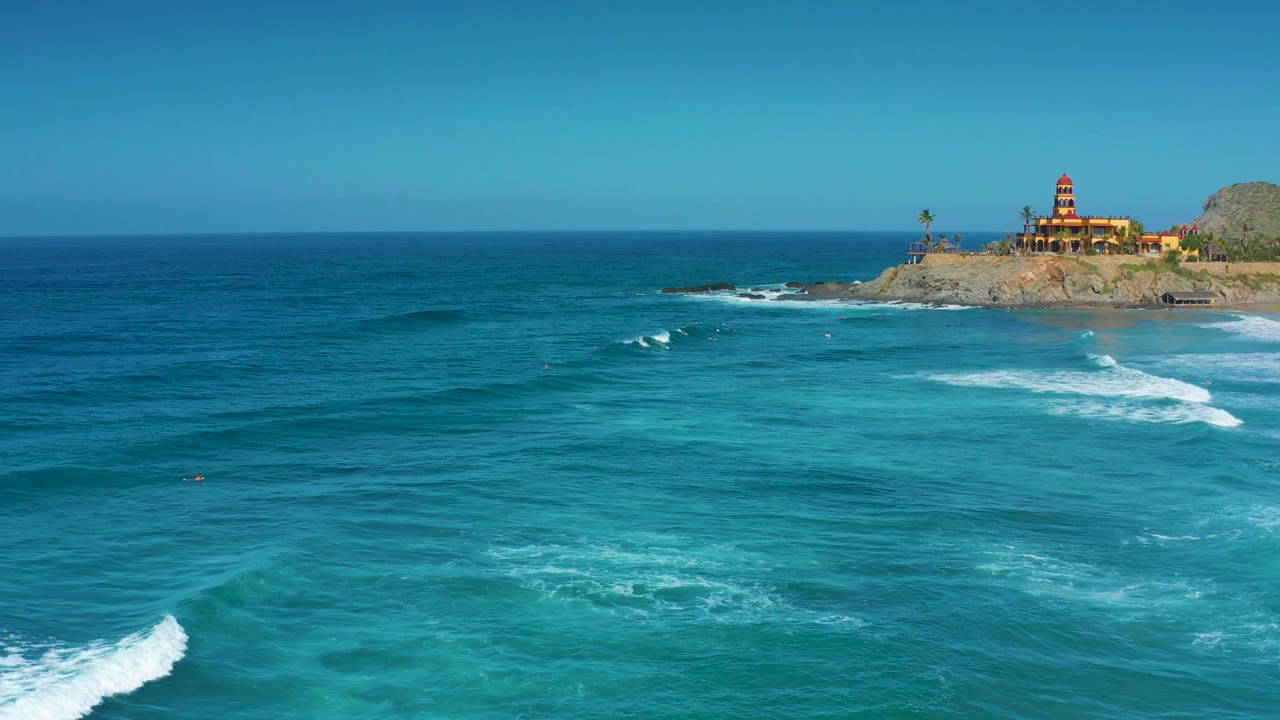 Aerial coastal view in Cerritos, Baja California Sur with darker blue water near the horizon, pale turquoise shallows, small white wave crests, and a building on the rocky point.