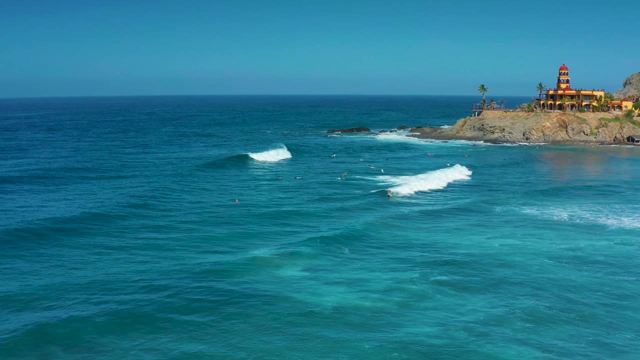 At 0:01 in Cerritos, Baja California Sur, white surf advances across the midground with the fort-like building still high on the rocky right edge and no visible people.