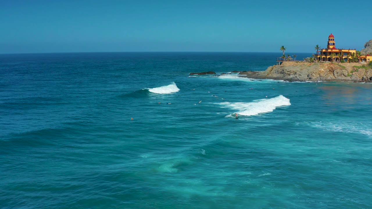 At 0:02 in Cerritos, Baja California Sur, the white wave line is farther right across the blue water, with the fort-like building cropped near the upper-right corner and no visible people.