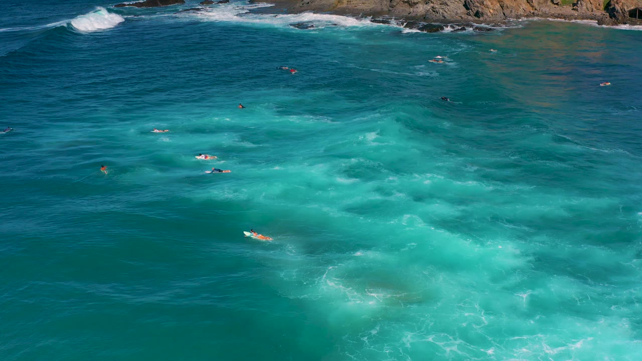 High aerial over Cerritos water with a broad turquoise section, several tiny board riders, scattered foam, and rocks visible across the upper frame.