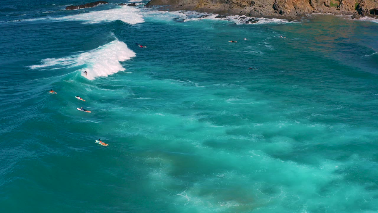 High aerial view over Cerritos, Baja California Sur, with turquoise water, a white breaking wave near the upper left, rocky shoreline at the top, and small distant surfers with no recognizable faces.