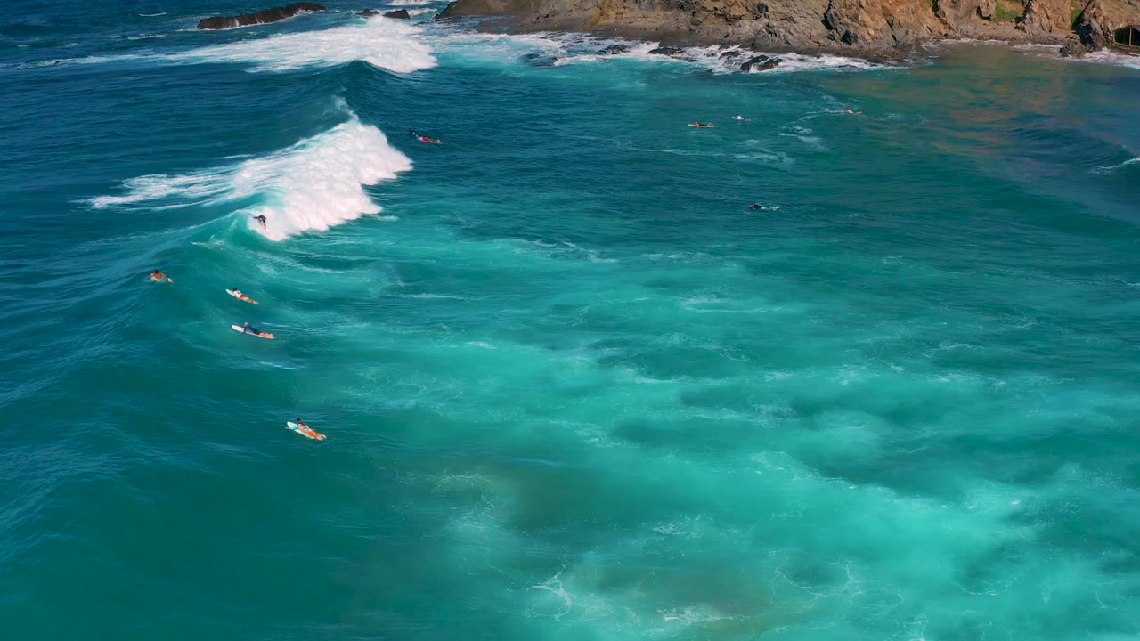 Aerial frame over Cerritos showing a white wave curling across blue-green water, several small surfboards below the foam line, rocky coast along the top, and no recognizable faces.