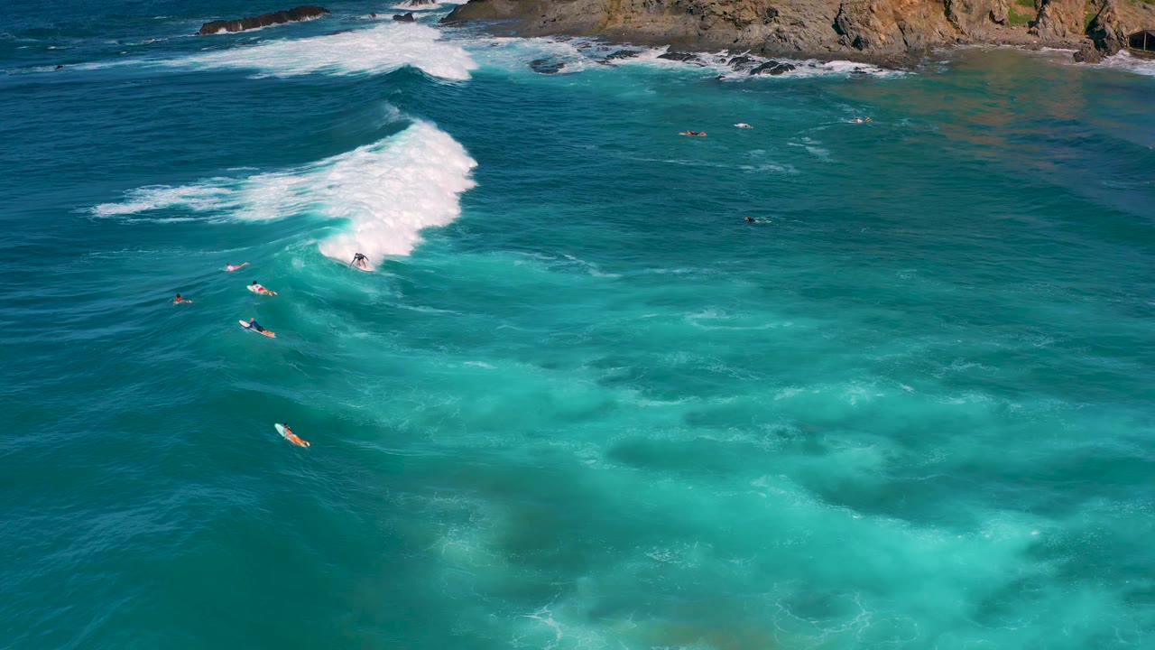 Cerritos aerial view with the white surf mass farther across the frame, scattered surfers and boards in the surrounding water, pale sand-colored shallows below, and no recognizable faces.