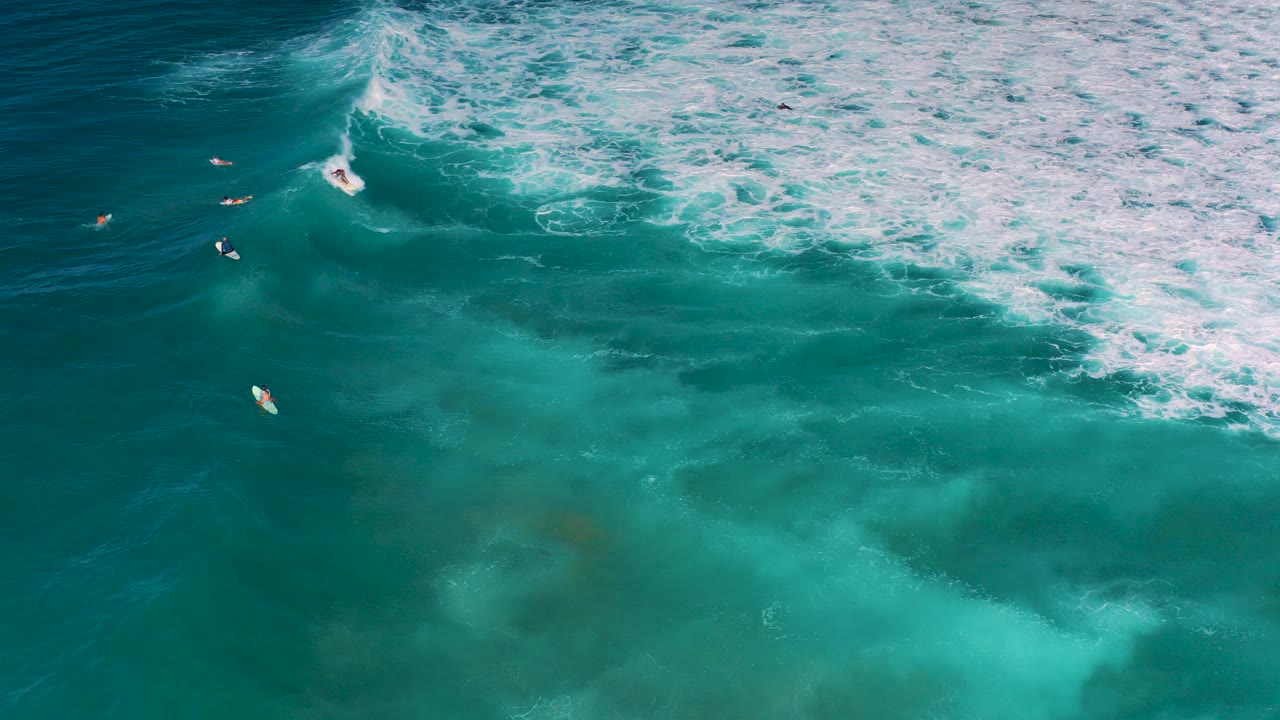 Overhead frame from Cerritos, Baja California Sur showing a surfer beginning to ride a white breaking wave while other surfers wait in blue water nearby.