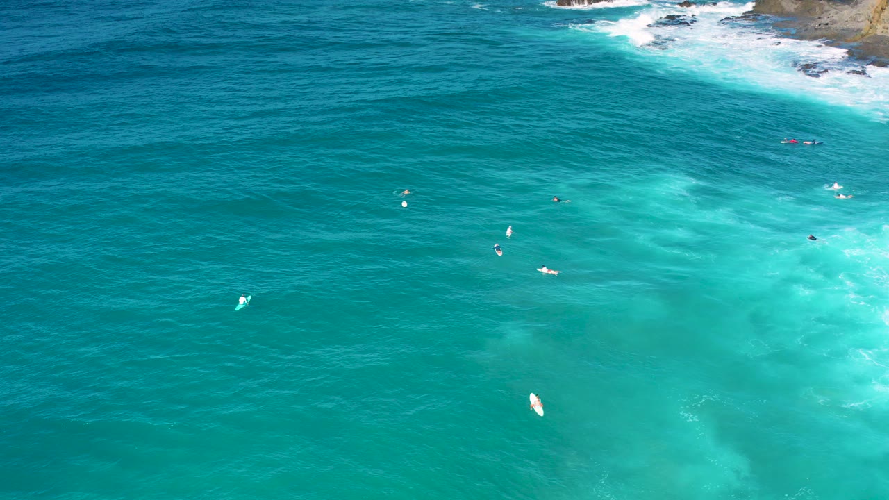 Aerial frame over Cerritos, Baja California Sur, showing turquoise water, white wave wash along rocks at the top edge, scattered small boards, and no visible people.
